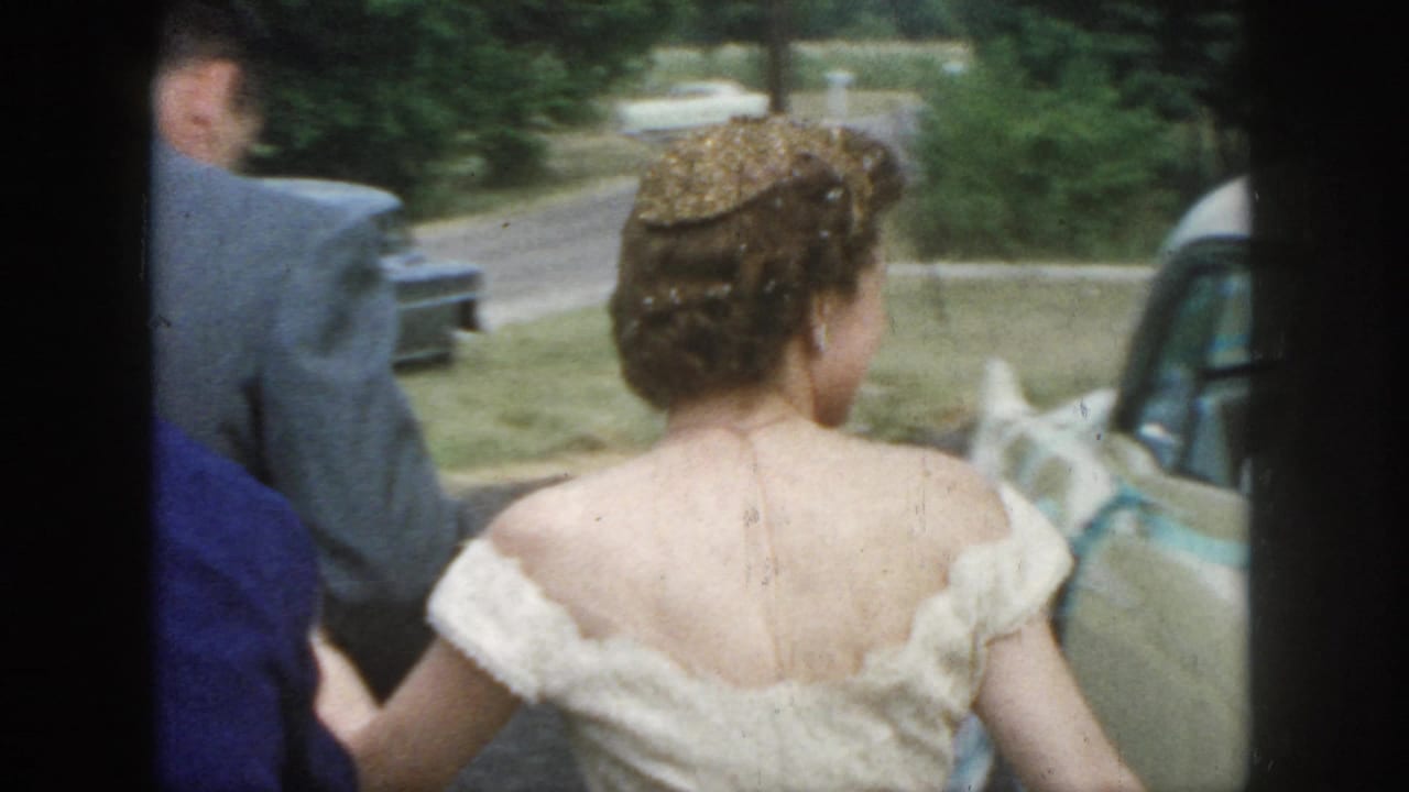 Bride seen from behind walking beside a suited man near a parked car.