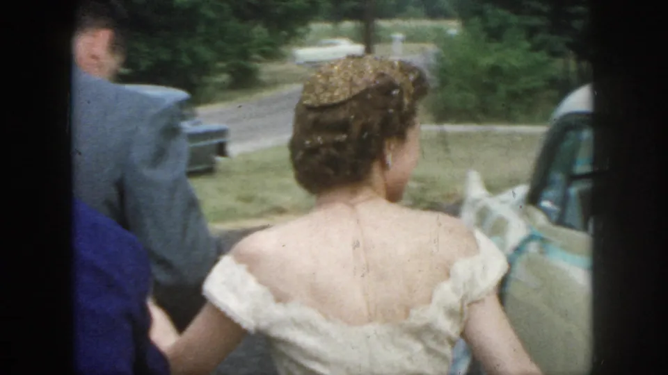 1958 Dayton wedding footage shows the bride from behind in an off-the-shoulder dress beside a suited man and parked cars.