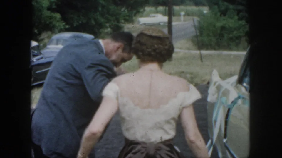 1958 Dayton home-movie frame of the bride and suited man beside a car, with another mid-century automobile visible nearby.
