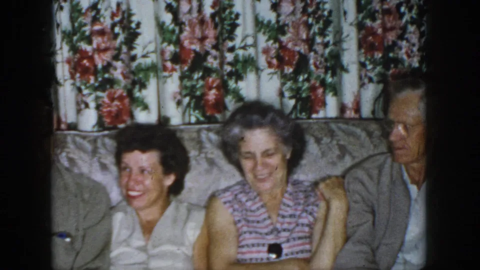 1958 Dayton indoor home-movie frame showing smiling family members seated on a couch before floral-patterned curtains.