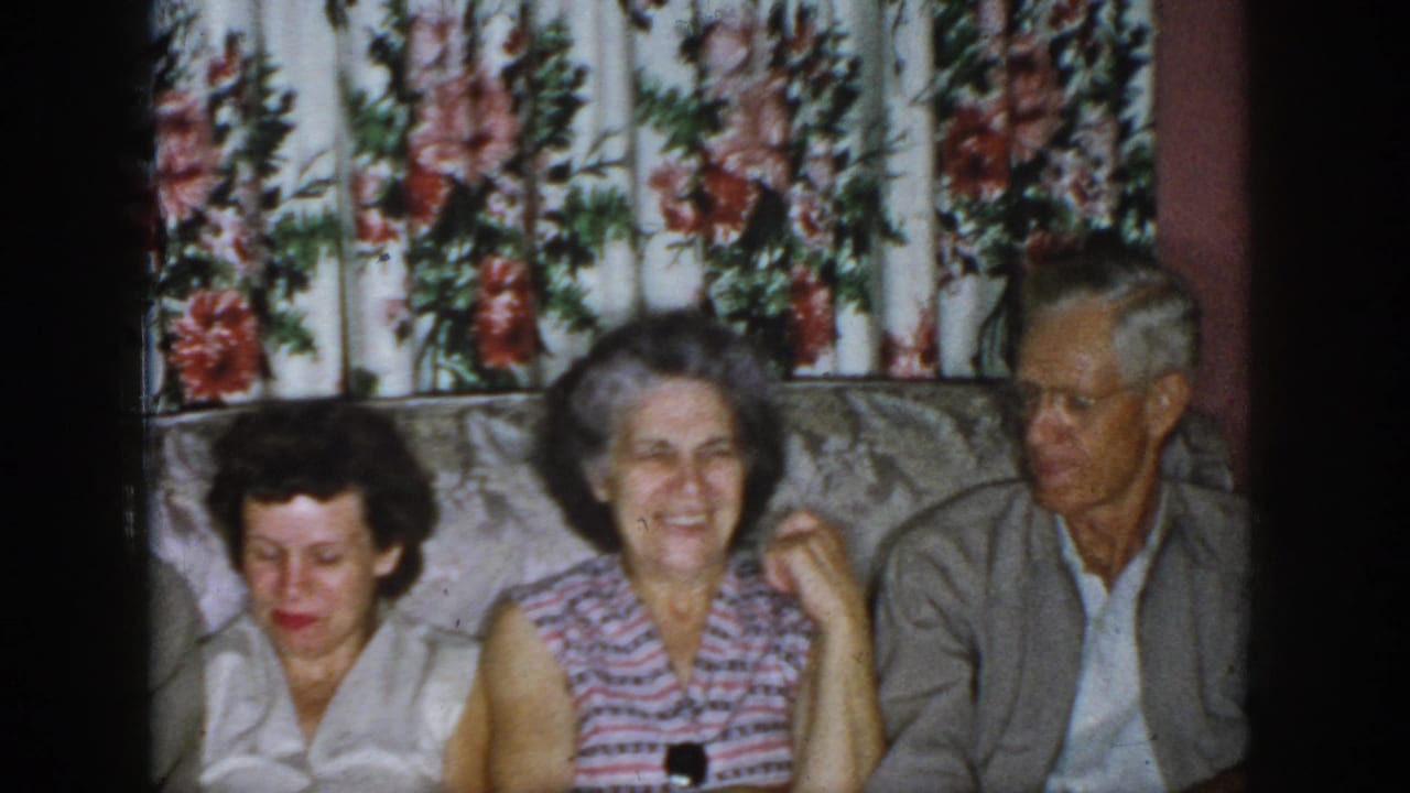 1958 Dayton home-movie frame showing three adults seated on a couch before floral curtains in a domestic living room.