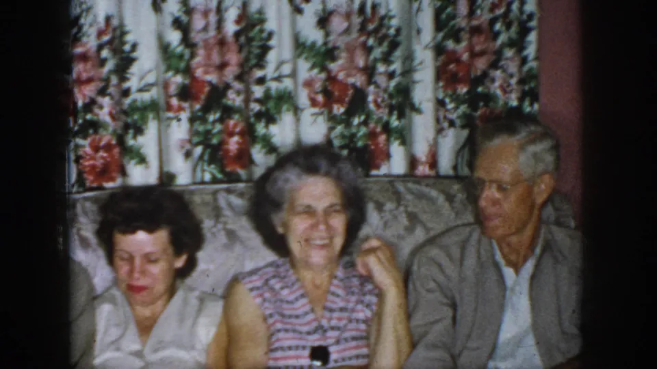 1958 Dayton home-movie frame showing three adults seated on a couch before floral curtains in a domestic living room.