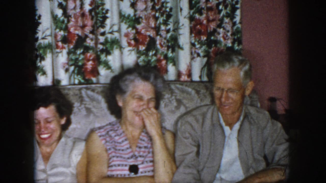 Three adults laugh on a couch, with the older woman covering her mouth and floral curtains behind them.