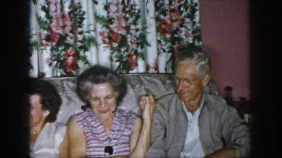 1958 Dayton home-movie frame showing adults seated in a living room, with floral curtains and couch upholstery visible.