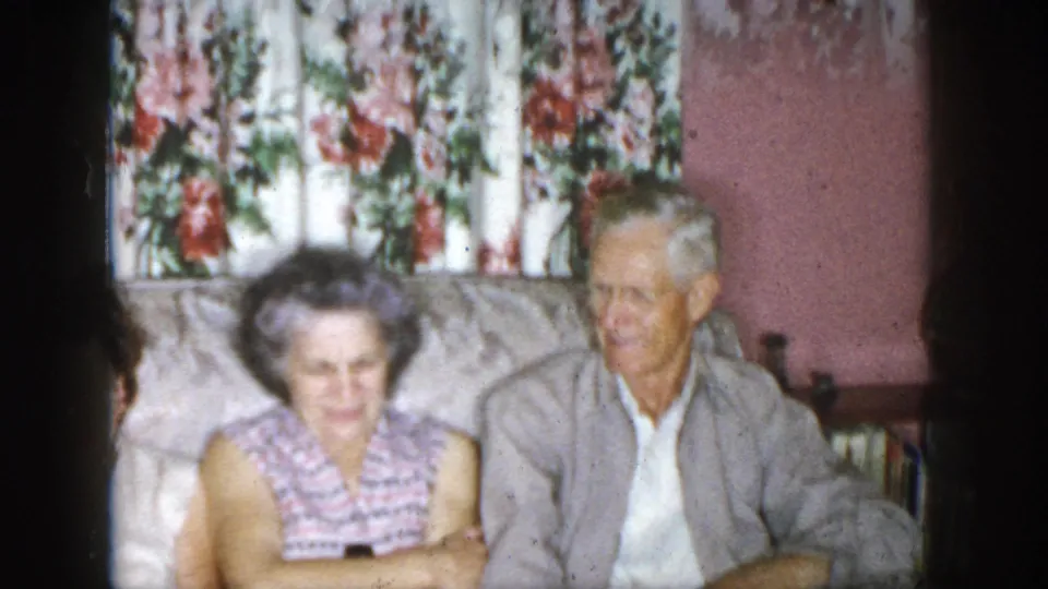 1958 Dayton home-movie frame focused on an older woman and older man seated on a couch in a floral-curtained living room.