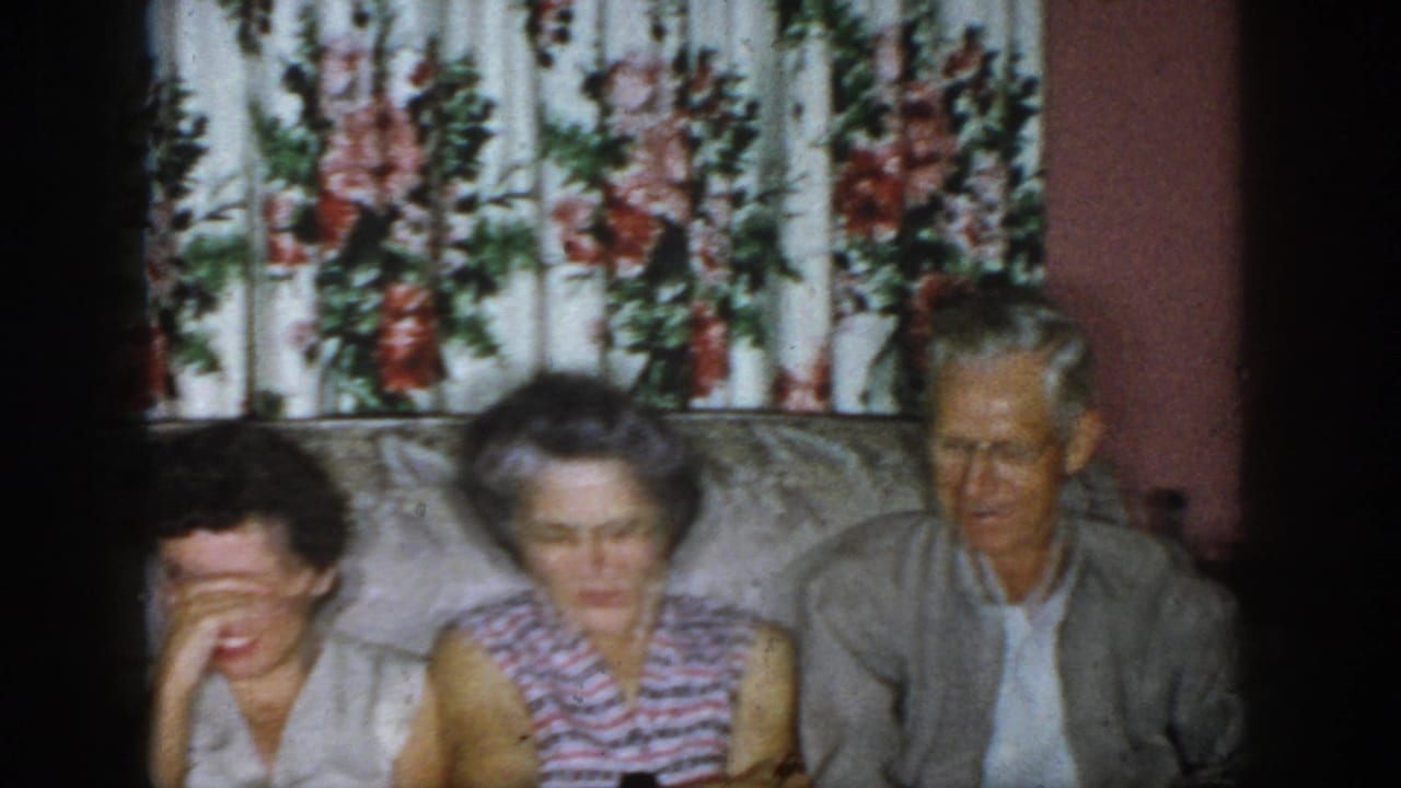 1958 Dayton home-movie frame showing adults seated closely on a floral couch in a private living-room interior.