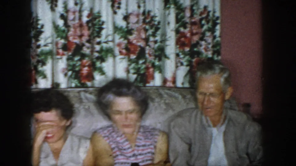 1958 Dayton home-movie frame showing adults seated closely on a floral couch in a private living-room interior.