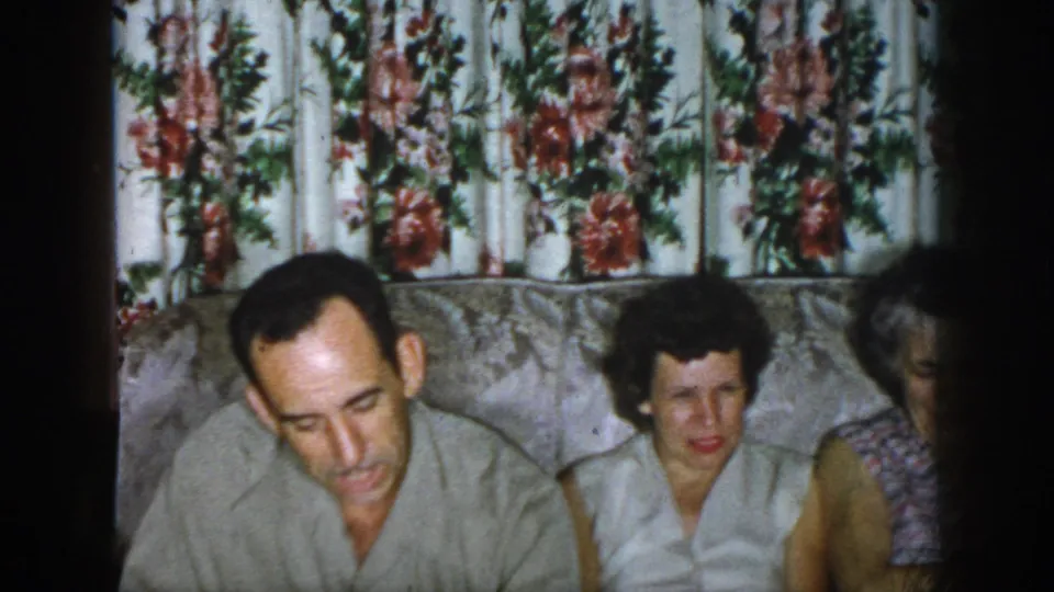 1958 Dayton home-movie frame showing a man leaning forward with two women seated beside him in a living room.
