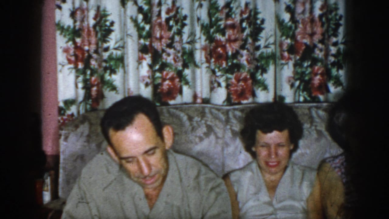 1958 Dayton, Ohio home-movie frame showing a man and woman seated indoors on a patterned sofa beneath floral curtains.