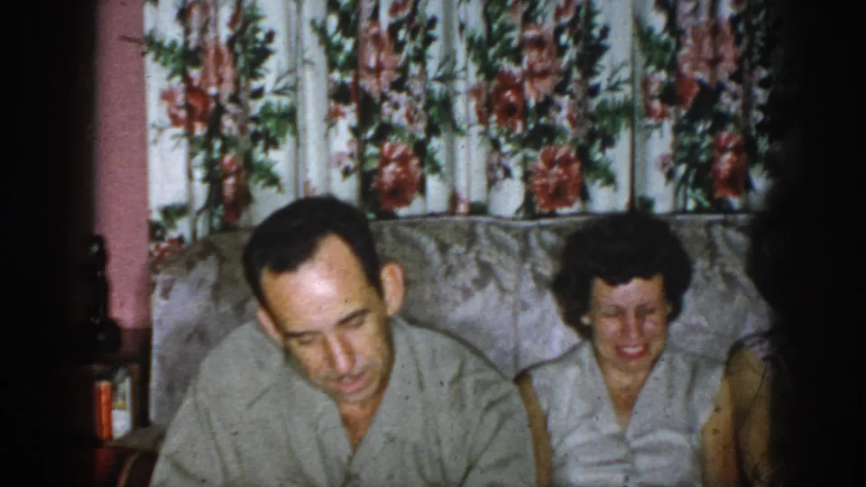 1958 Dayton, Ohio interior frame with a man leaning forward, a smiling woman on the sofa, and another adult partly visible at right.