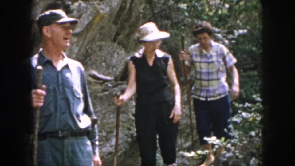 Three hikers pause on a rocky wooded trail, with hats and walking sticks visible in the 1958 Dayton footage.