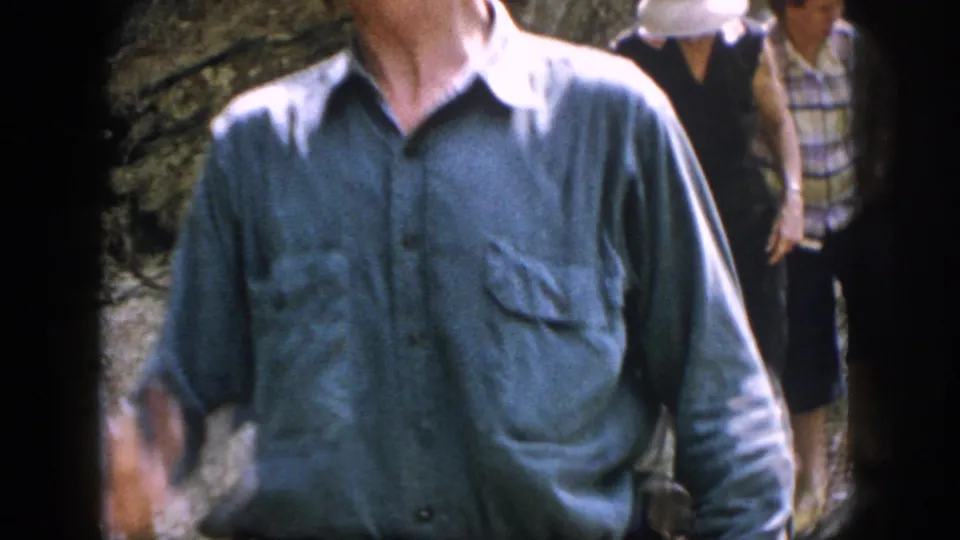 A close handheld view shows the older man crossing in front while two companions follow over rocks.