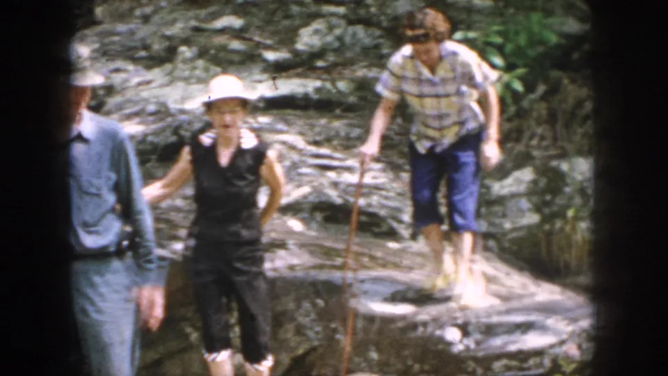 The group spreads out while crossing wet-looking rocks or a shallow rocky area in wooded Dayton, Ohio footage from 1958.