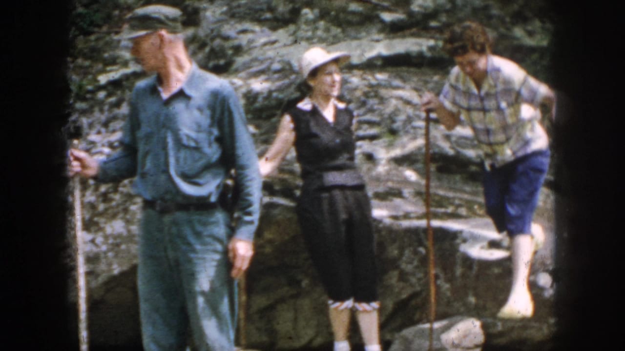 1958 Dayton home-movie frame showing three adults navigating rocky terrain with walking sticks during an outdoor outing.