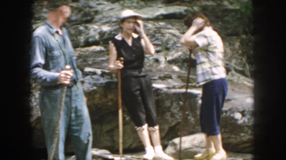 1958 Dayton home-movie frame showing adults pausing on rocky ground, with walking sticks and casual summer clothing visible.