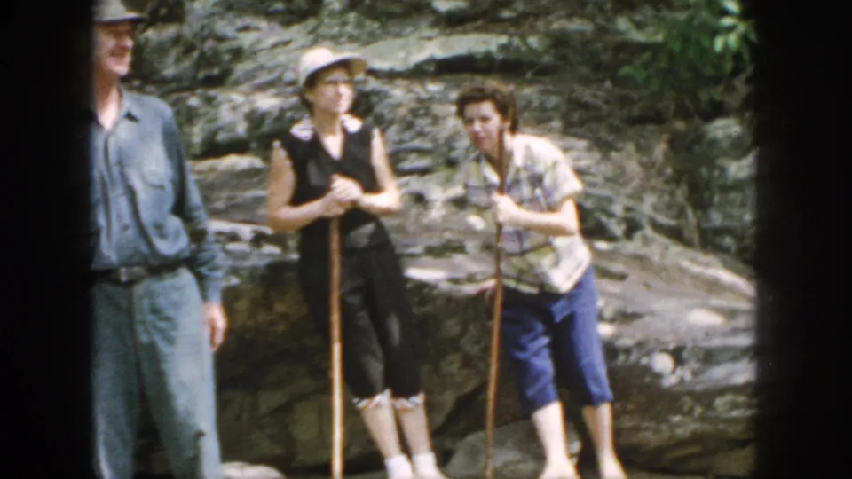 1958 Dayton home-movie frame of three adults regrouping on uneven rocks during a casual hiking outing.