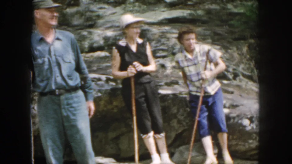 In metadata-supported 1958 Dayton, Ohio footage, three adult hikers pause on rocky terrain with walking sticks near a stream.