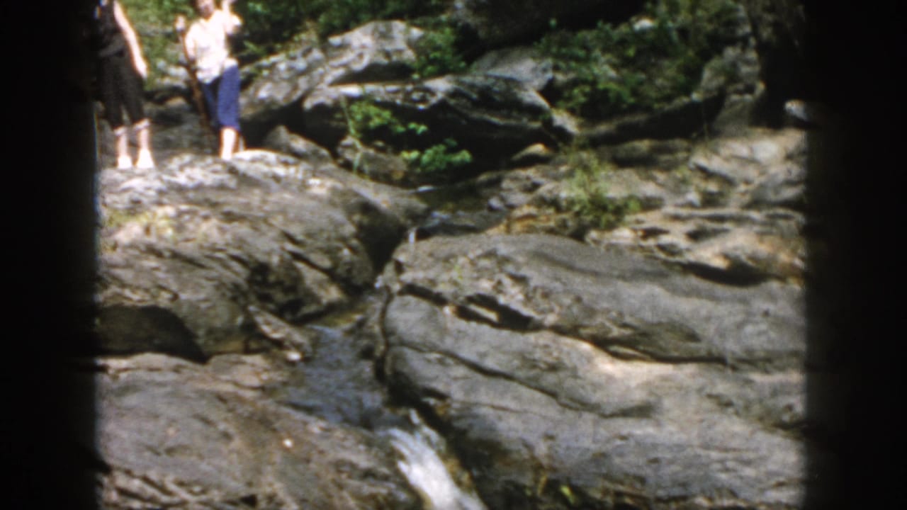 1958 Dayton, Ohio home-movie frame showing adults on gray rocks near a small wooded stream and waterfall.