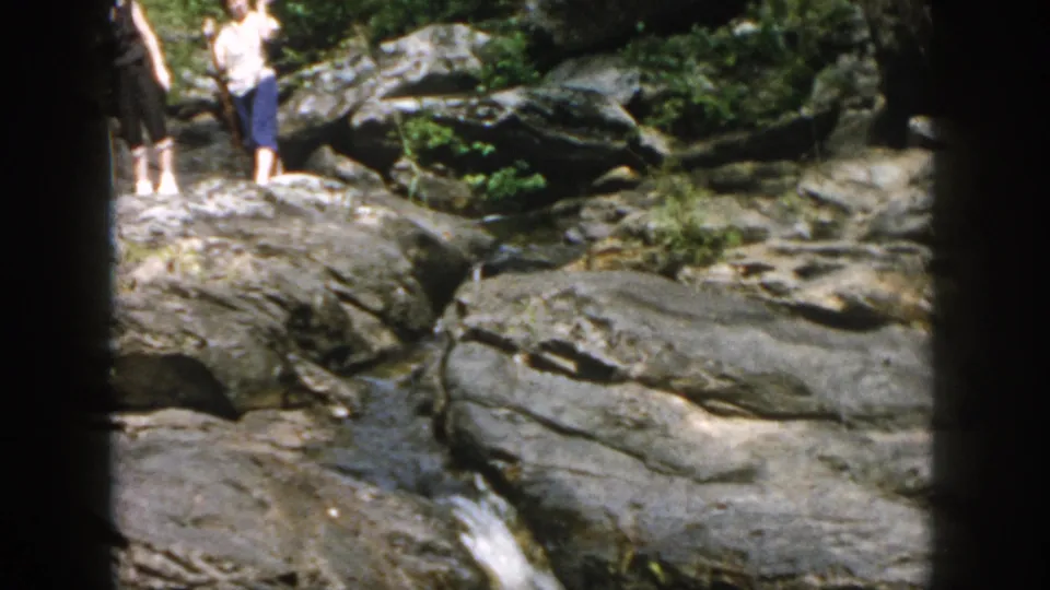 1958 Dayton, Ohio home-movie frame showing adults on gray rocks near a small wooded stream and waterfall.