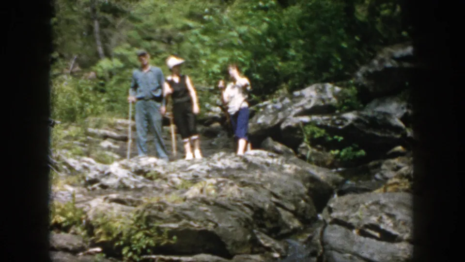 A home-movie view of three adults on rocks by a stream in Dayton, Ohio, with walking sticks and summer foliage.