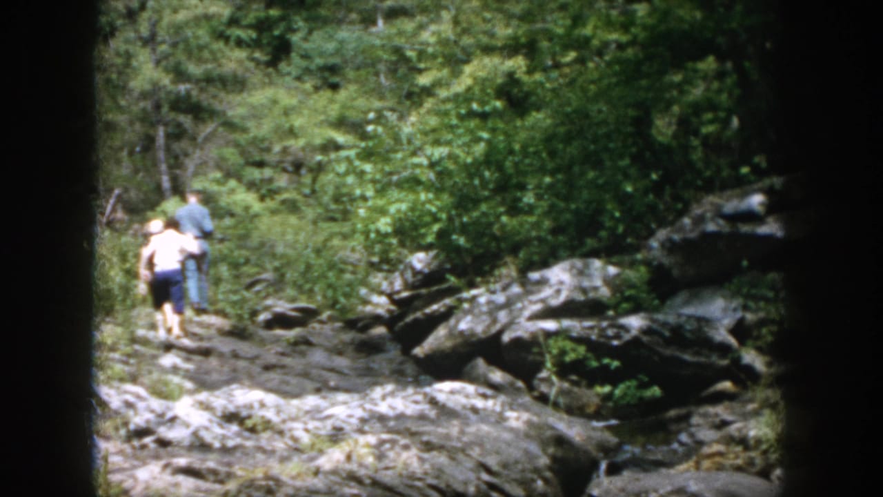 Two hikers move along a rocky woodland path in 1958 Dayton, Ohio home-movie footage, seen from a distant 8mm perspective.