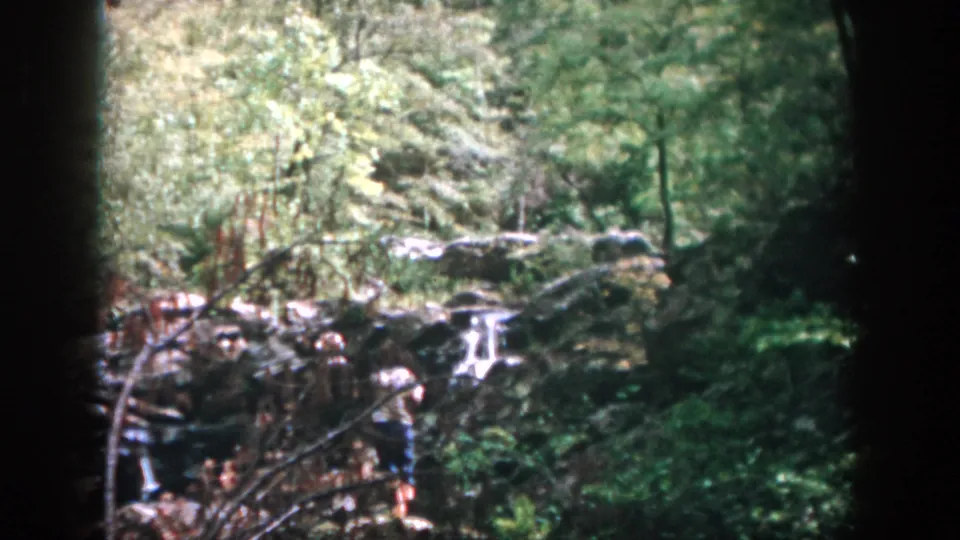 A 1958 Dayton home-movie frame shows a small rocky waterfall in the woods, with a person partly visible at the right edge.