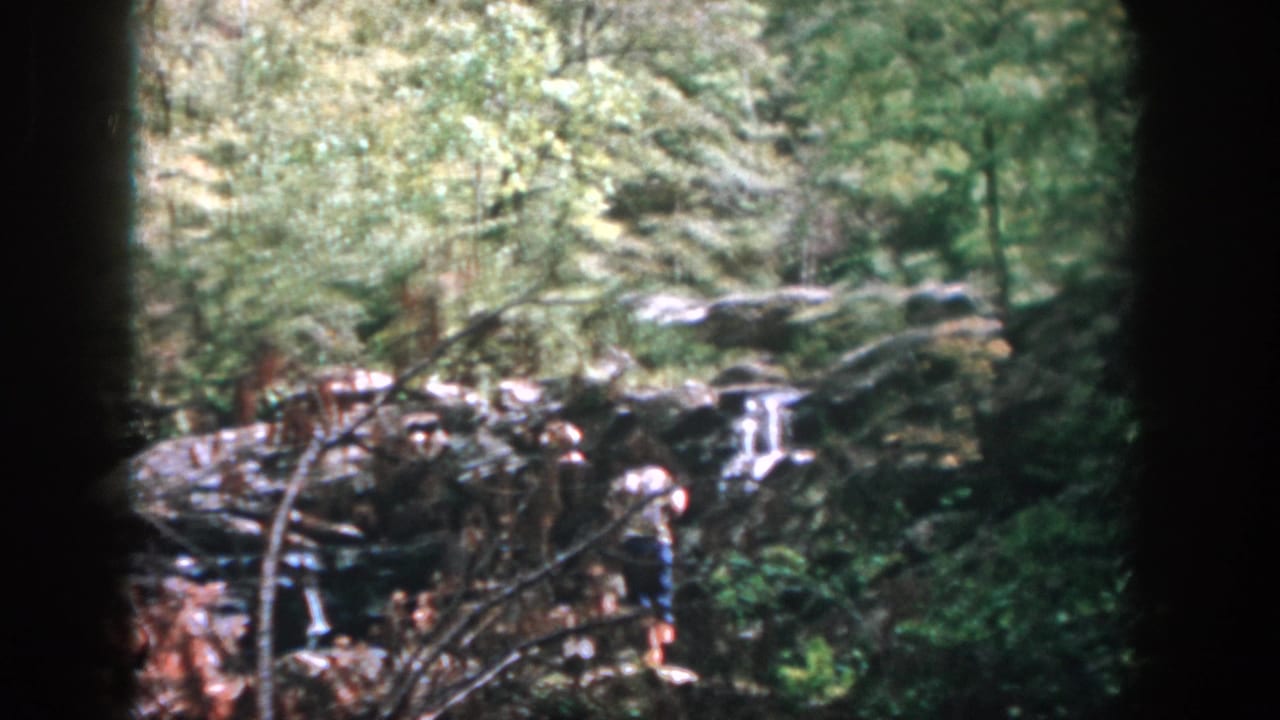 1958 Dayton-area home-movie frame showing two people on rocky ground beside a small waterfall in dense woods.