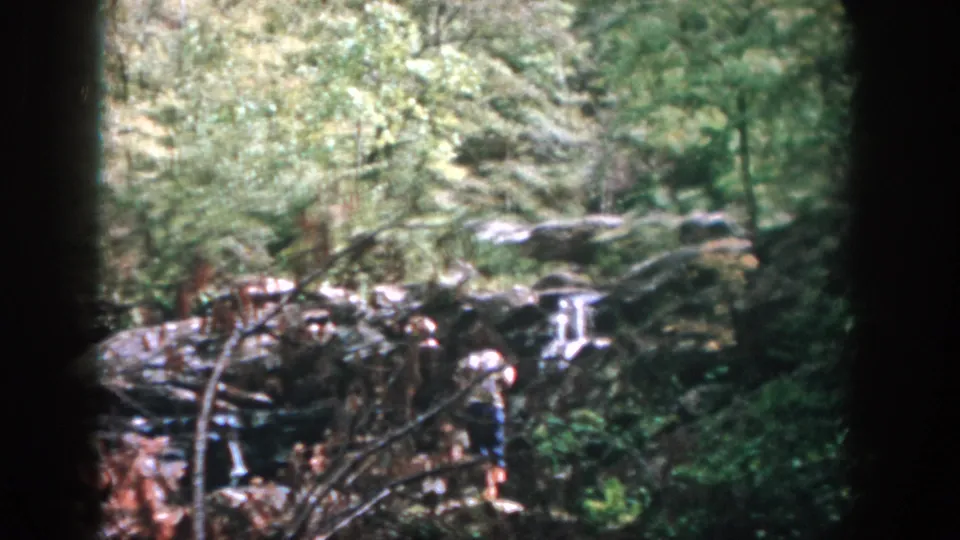 1958 Dayton-area home-movie frame showing two people on rocky ground beside a small waterfall in dense woods.