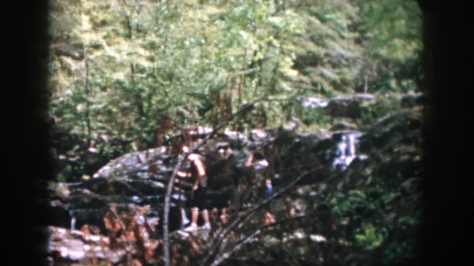 A handheld 8mm frame shows two people partly hidden by branches as they move through rocky woods near water.