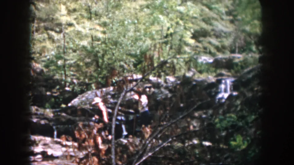 The final frame shows two distant visitors near rocks and a small waterfall, framed by trees and dark 8mm film edges.