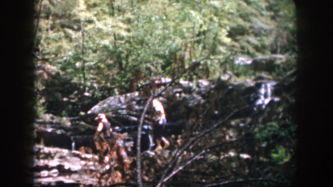 In supported 1958 Dayton home-movie footage, two adults step across jagged creek rocks with forest growth and small falls behind them.