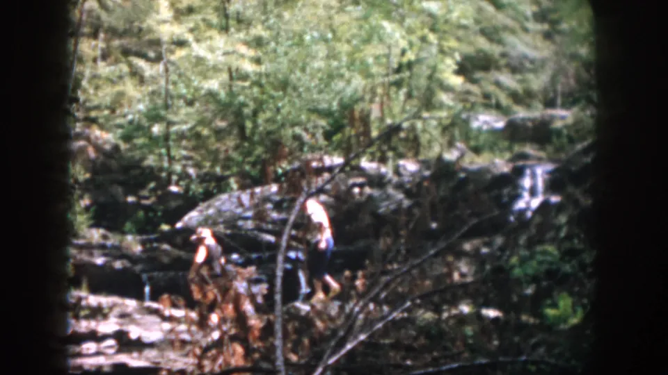 In supported 1958 Dayton home-movie footage, two adults step across jagged creek rocks with forest growth and small falls behind them.