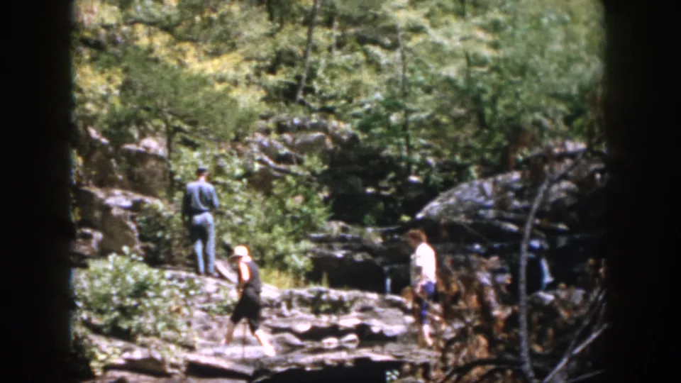 The final frame shows three adults in the supported 1958 Dayton creek footage, spaced across rocks with water, branches, and forest vegetation around them.