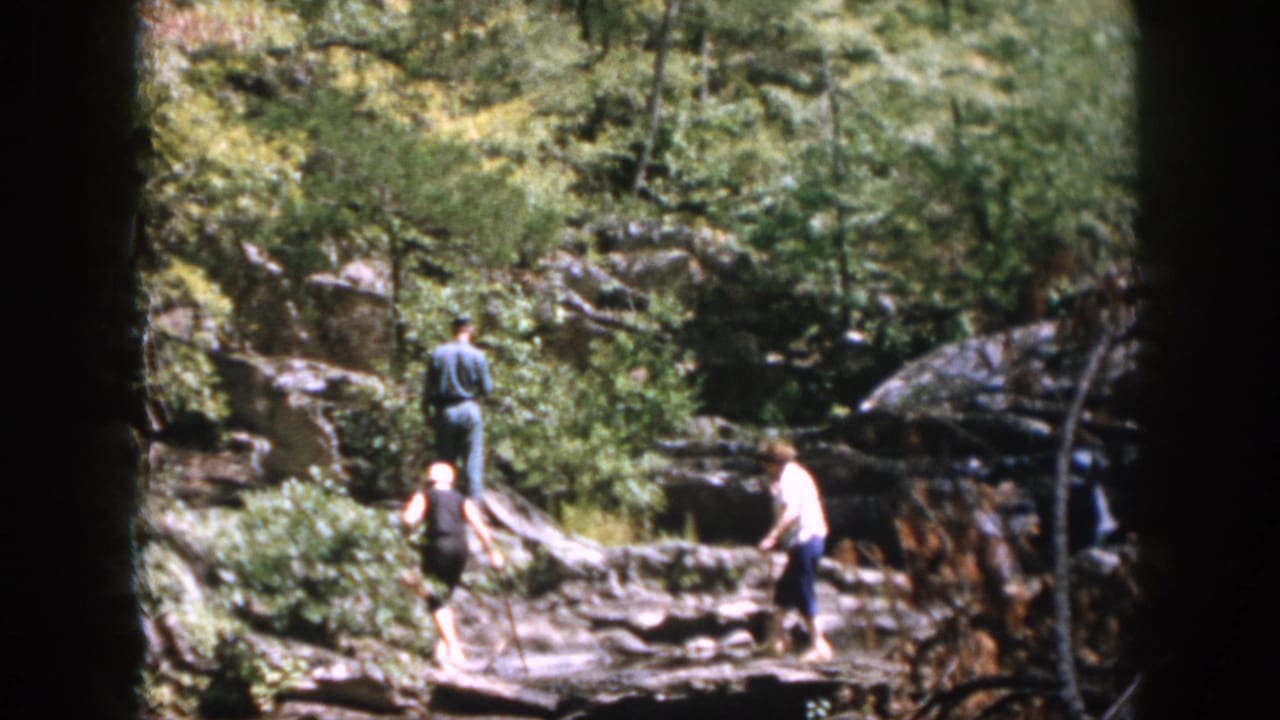 In the supplied 1958 Dayton home-movie frame, young people move through a rocky wooded ravine in summer light.