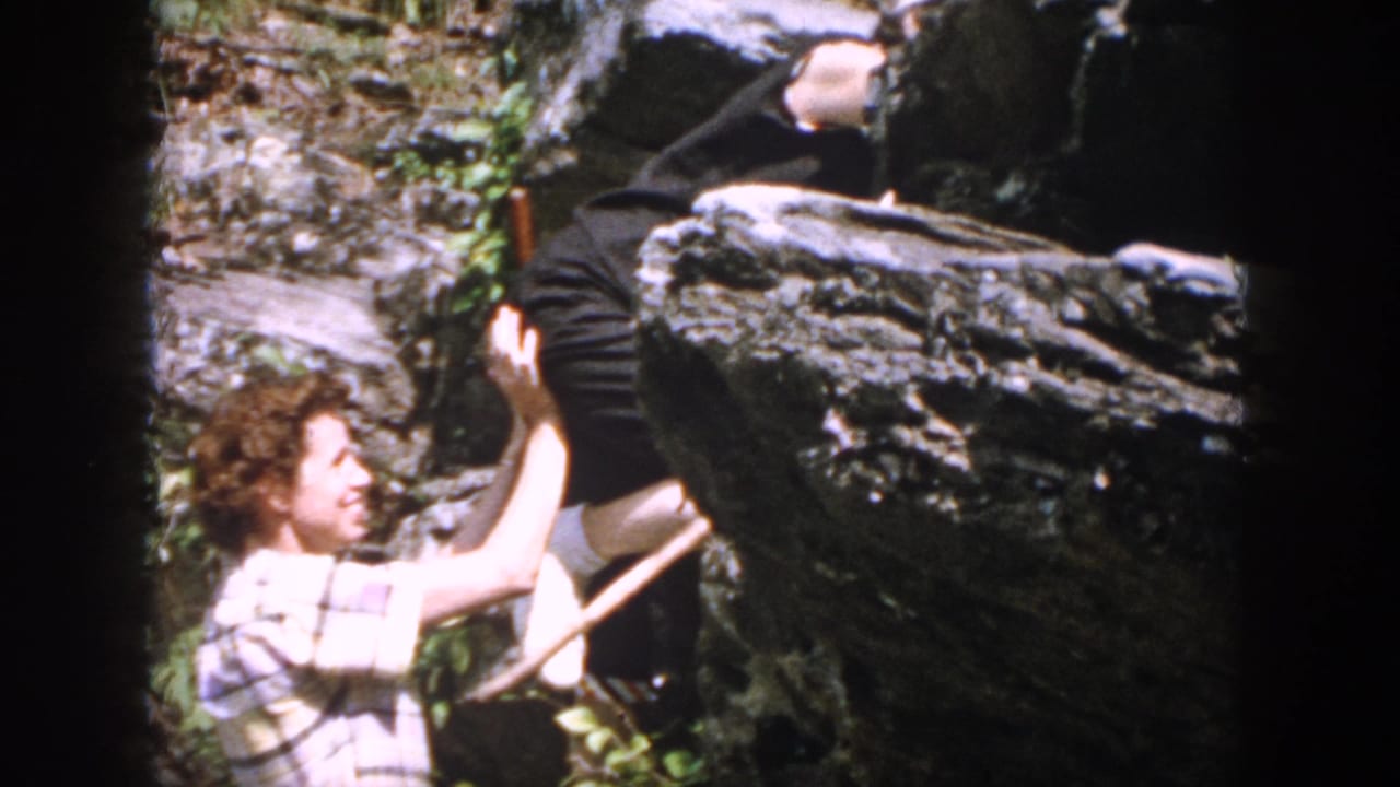 A smiling young man in a plaid shirt helps another person climb over a large rock.