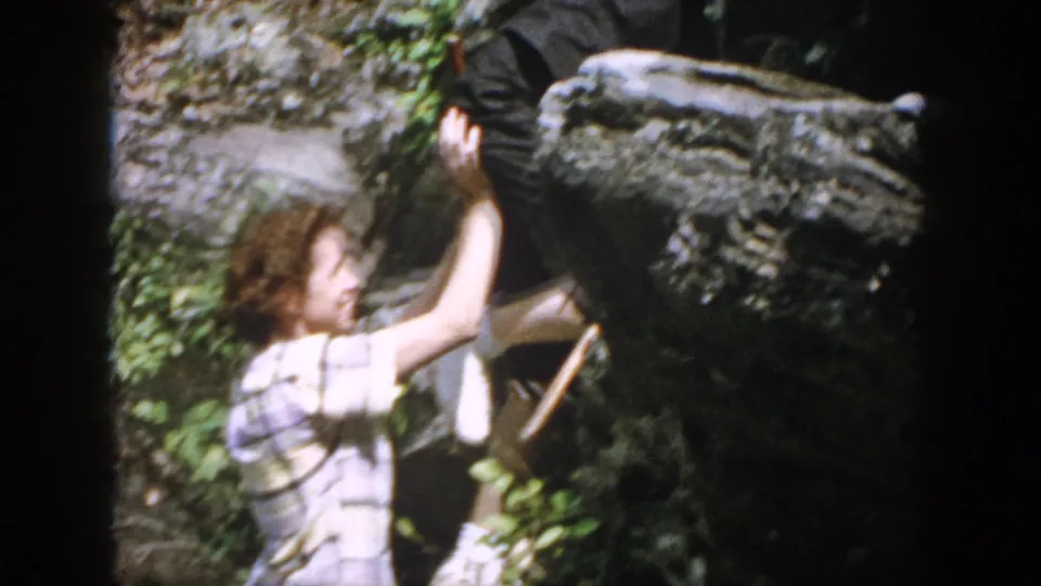 The home-movie frame shows close assistance during a rock scramble, with the helper partly blurred by motion and foliage.