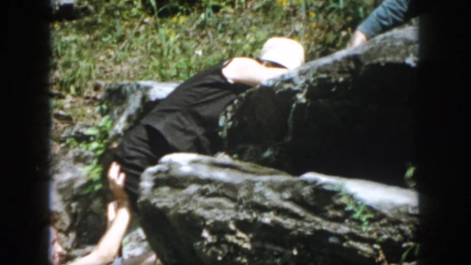 A climber in dark clothing crosses a boulder as another person supports them from below in the wooded ravine scene.