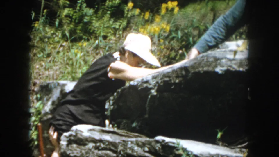 In 1958 Dayton-area home-movie footage, a woman in a white sun hat climbs among dark rocks and greenery.