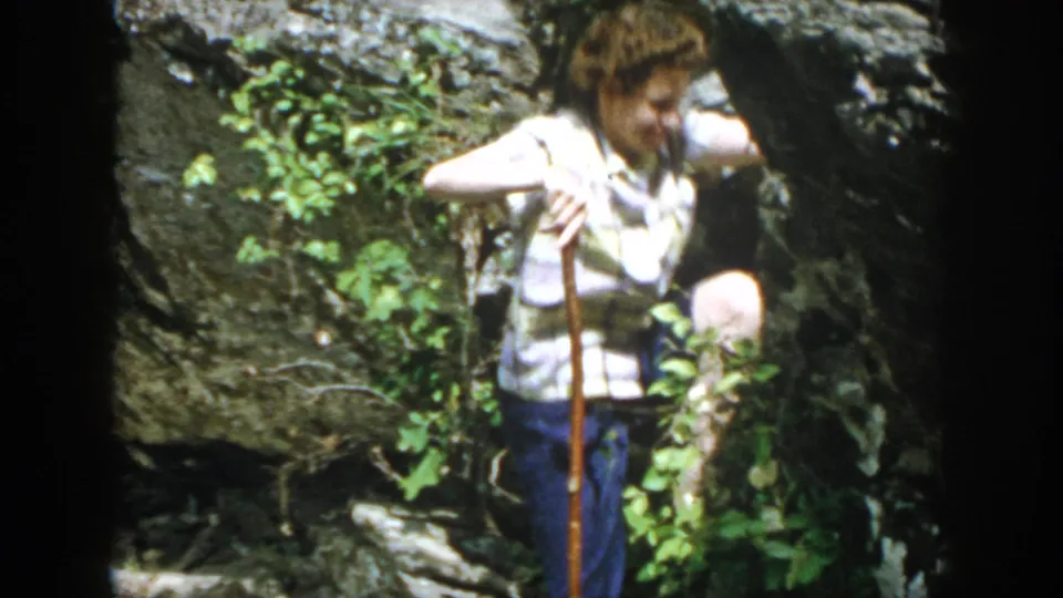 The woman, now lower in the rocky forest setting, holds a wooden cane beside greenery in the 1958 Dayton footage.