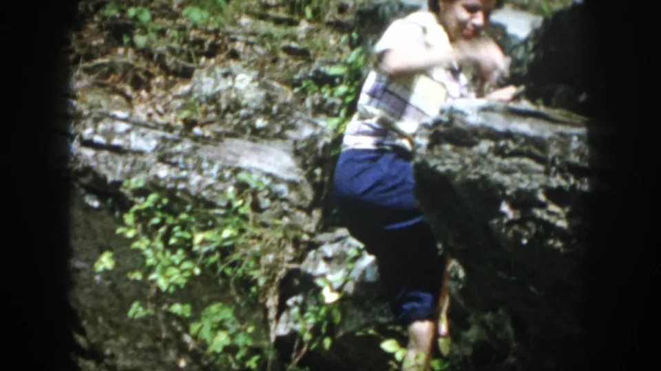 The woman continues moving along a rocky forest ledge, with her cane and surrounding foliage visible in the Dayton 1958 home movie.