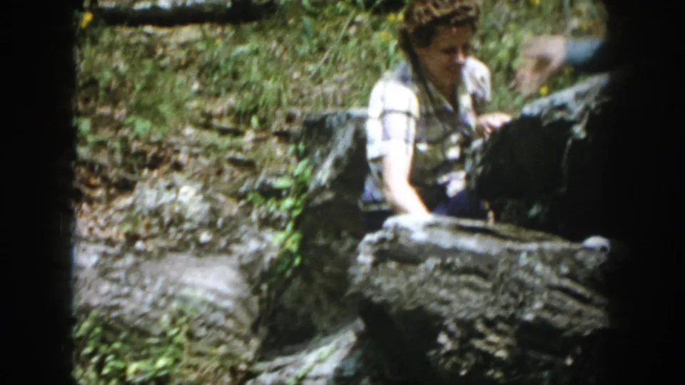 1958 Dayton, Ohio home-movie frame showing an adult in a plaid shirt moving among large rocks near foliage and water.