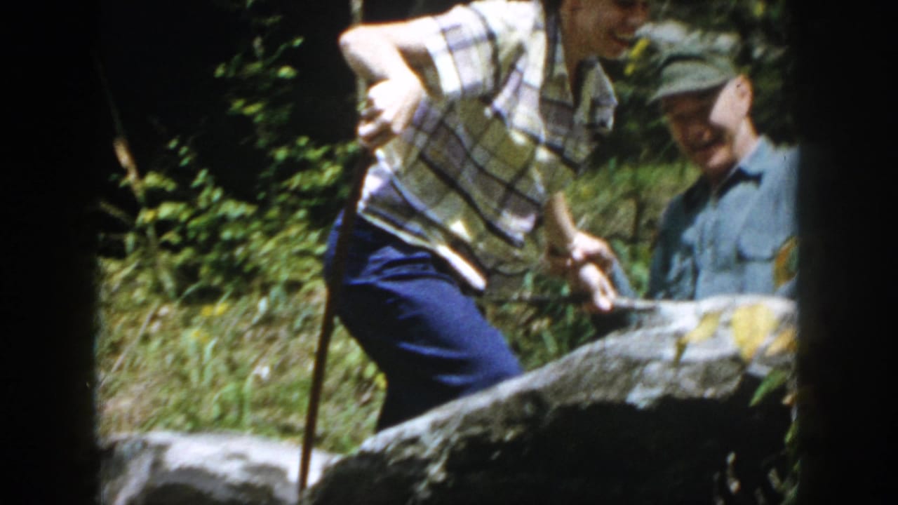 Two adults stand among rocks, one using a walking stick while another watches nearby.
