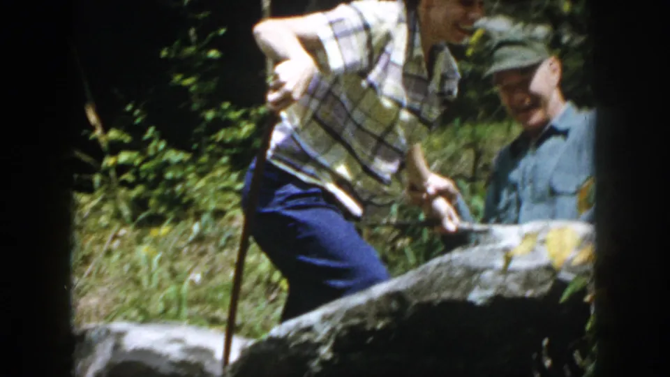 1958 Dayton, Ohio frame with two adults on rocks, one in a plaid shirt using a walking stick near wooded water.