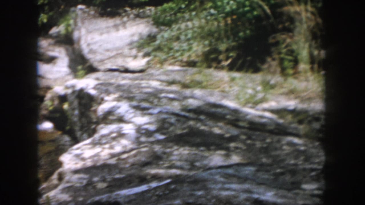 Large layered rocks beside a shaded stream with greenery above.