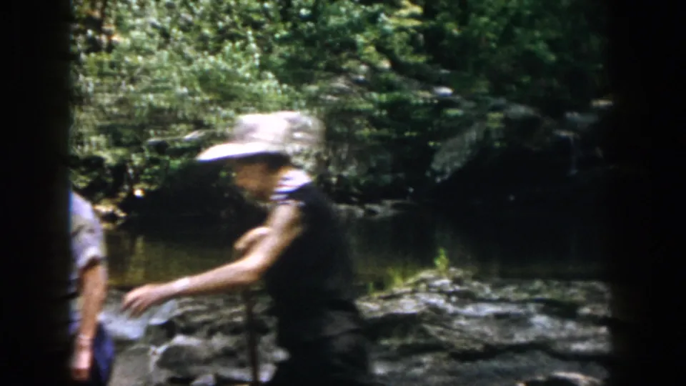 1958 Dayton-area home-movie frame showing an adult woman in a wide-brimmed hat crossing a shallow rocky stream.