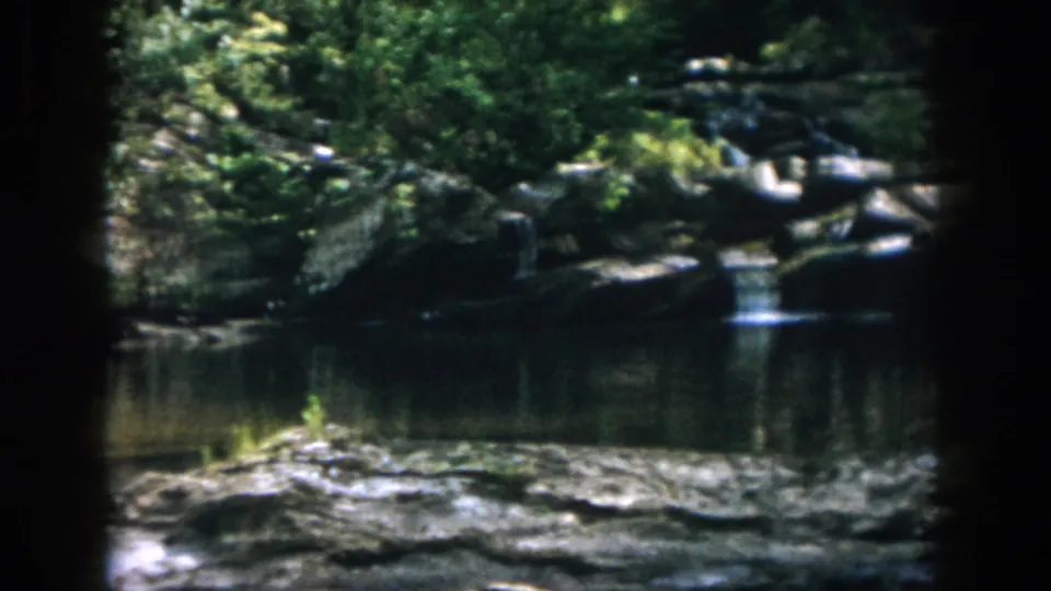 1958 Dayton-area frame of a quiet rocky stream, dense foliage, and small cascades in a wooded countryside setting.
