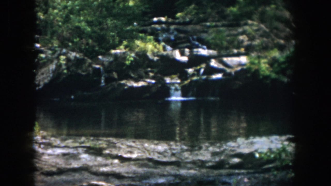 1958 8mm home-movie frame from Dayton, Ohio, showing a small waterfall, rocks, and still river water beneath dense trees.