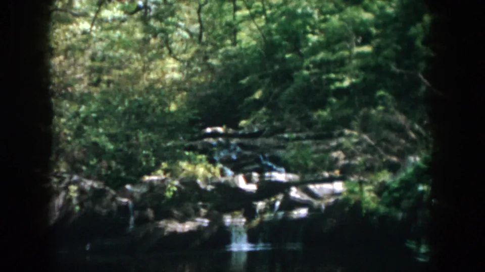 1958 8mm home-movie view of a wooded Dayton river scene, with a small cascade, rocks, and soft sunlight through trees.