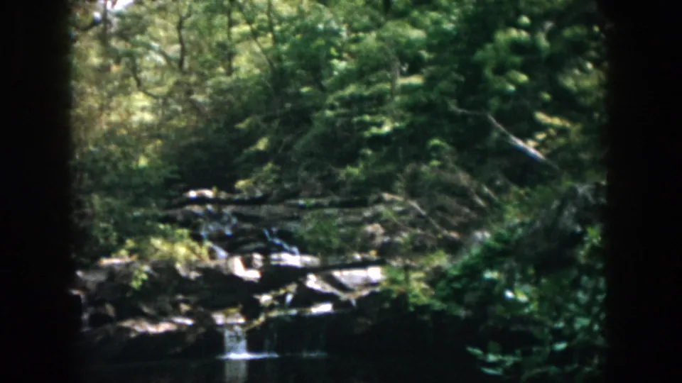 Final 1958 Dayton, Ohio frame showing a small waterfall, reflective river water, surrounding rocks, and dense wooded banks.