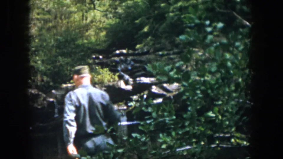 1958 Dayton-area 8mm frame with an adult man walking near a small pond and rocky waterfall in dense woods.